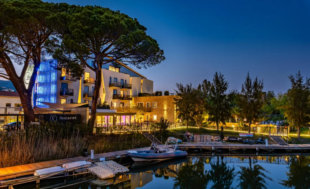 A modern building lit up with a restaurant on the ground floor, surrounded by trees and located near a body of water with moored boats.