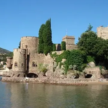 Medieval stone castle located by a body of water, surrounded by trees and vegetation, with a beach and people in the background.