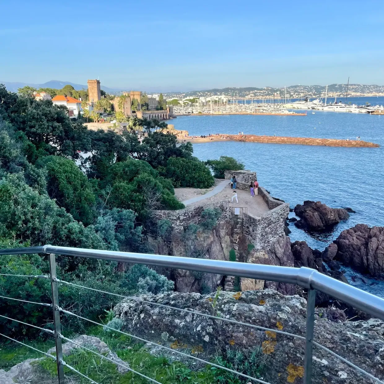 View of a marina with boats, a medieval fortress, and walkers on a coastal path