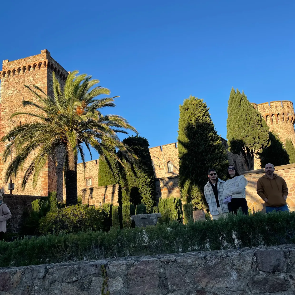 Four people pose in front of a stone castle surrounded by trees and vegetation.