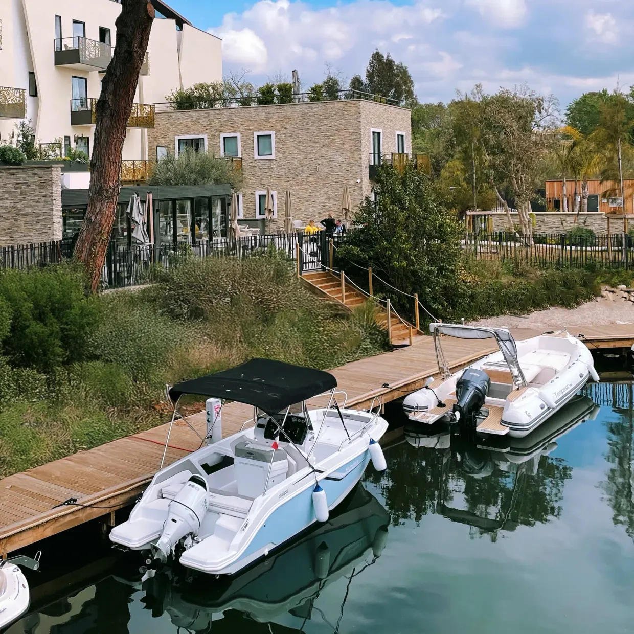 Wooden quays with several small motorboats moored in front of modern residential buildings surrounded by vegetation