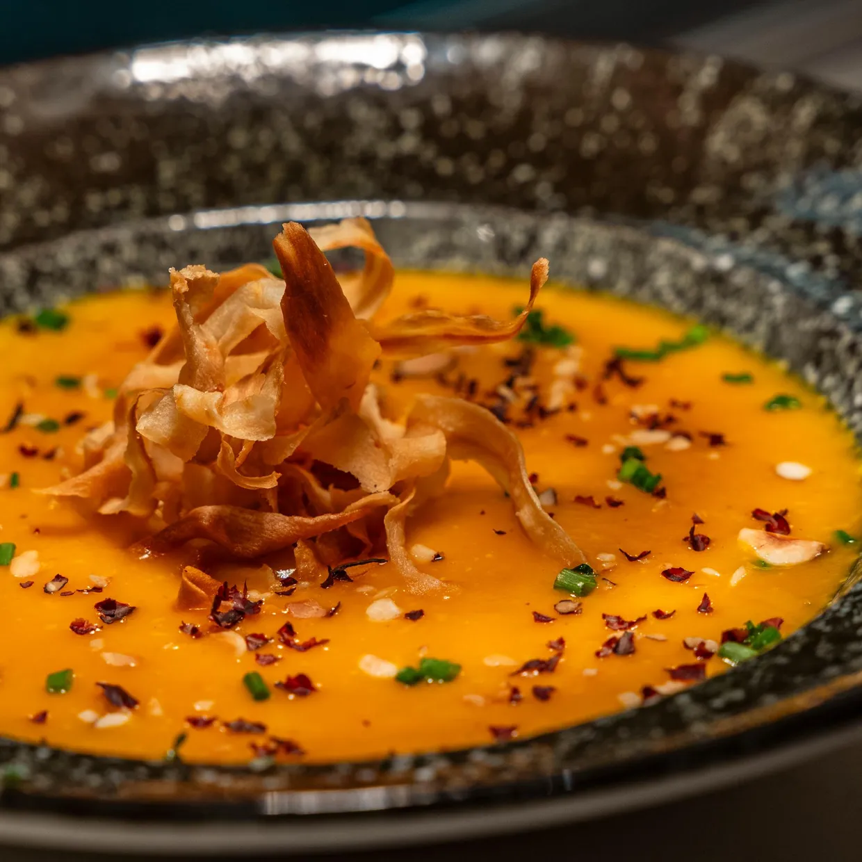 A bowl containing an orange soup topped with chips and fresh herbs, placed on a dark surface
