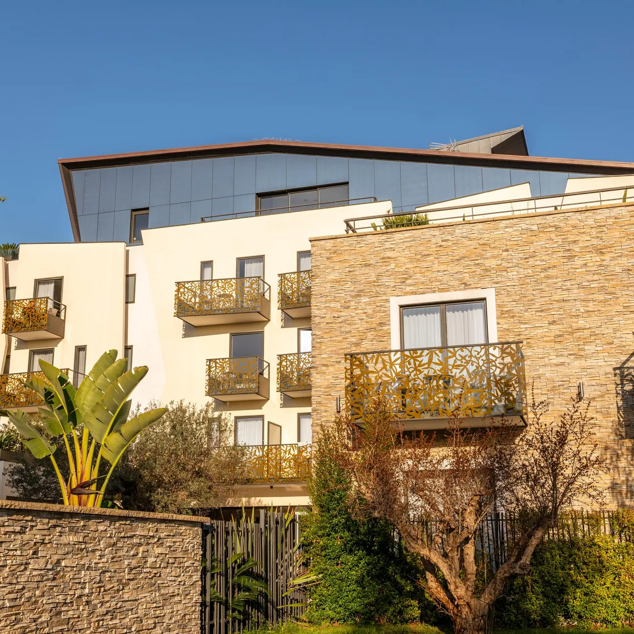 Modern building with balconies adorned with metal patterns and stone facade, surrounded by varied vegetation.