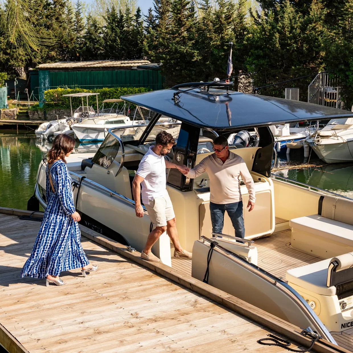 Three people board a boat moored in a port surrounded by other vessels and vegetation.