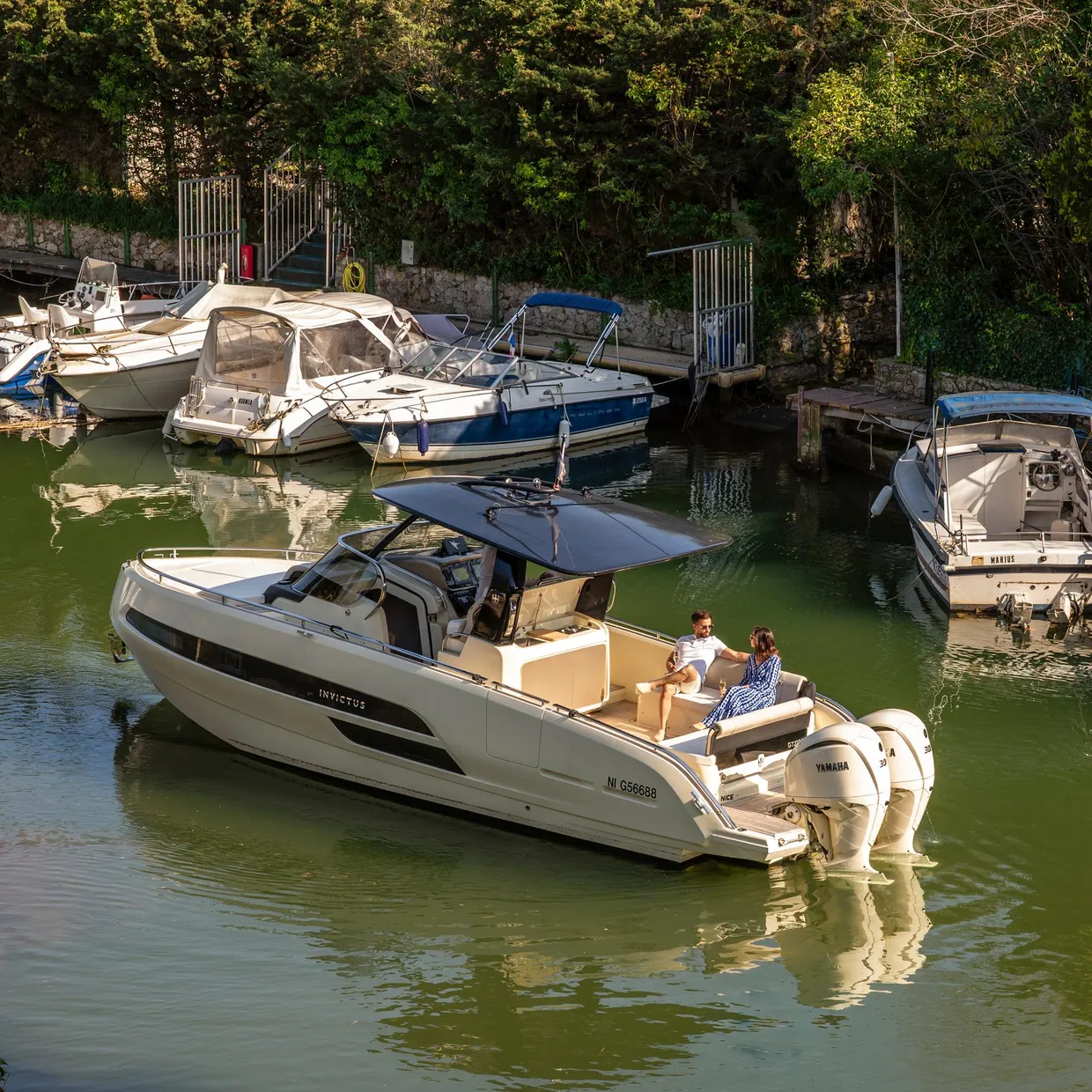 A white pleasure boat with two people on board is moored in a small port surrounded by other vessels and vegetation.