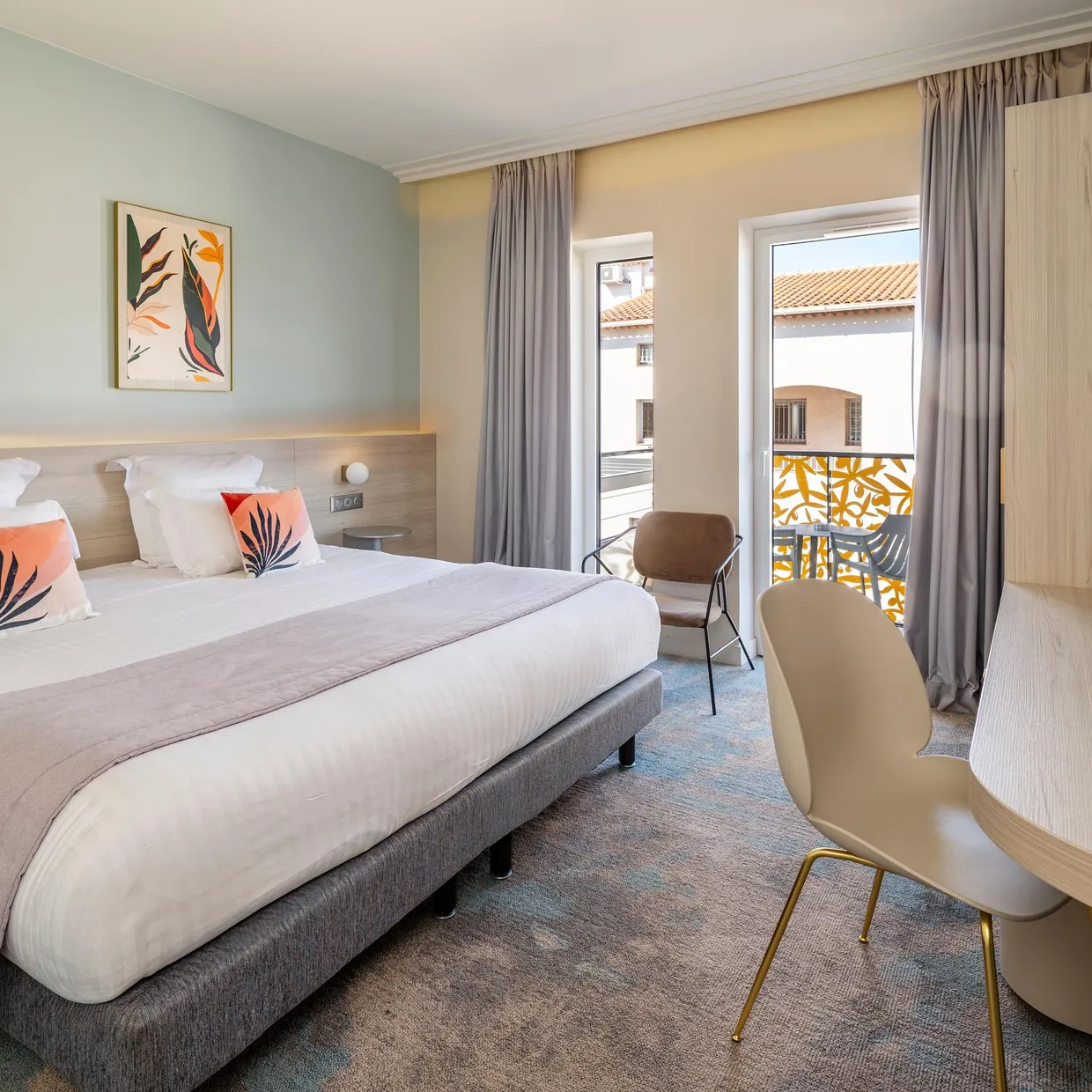 Hotel room with a double bed, a bedside table, a chair, and a French door leading to a balcony with a view of rooftops.