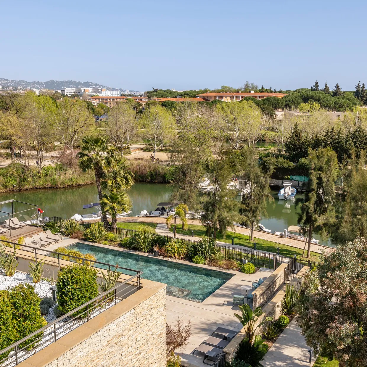 Aerial view of a garden with a pool, bordered by trees and a body of water where boats are moored.