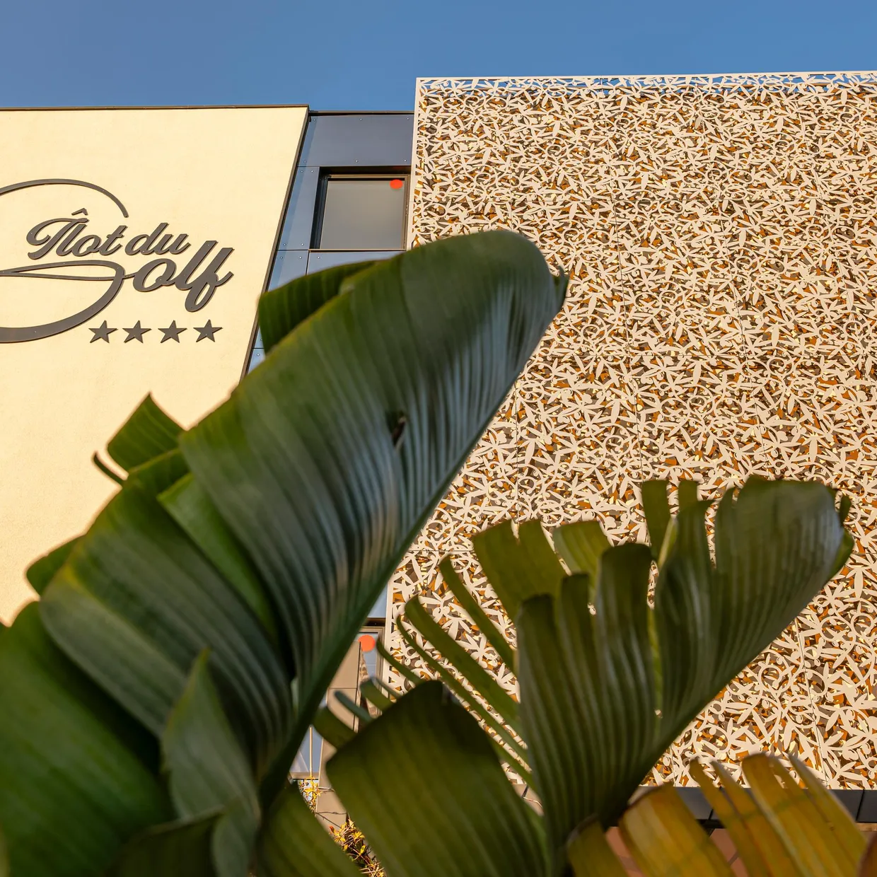 Facade of a building with the sign "Hôtel du Golf" and four stars, partially obscured by plant leaves