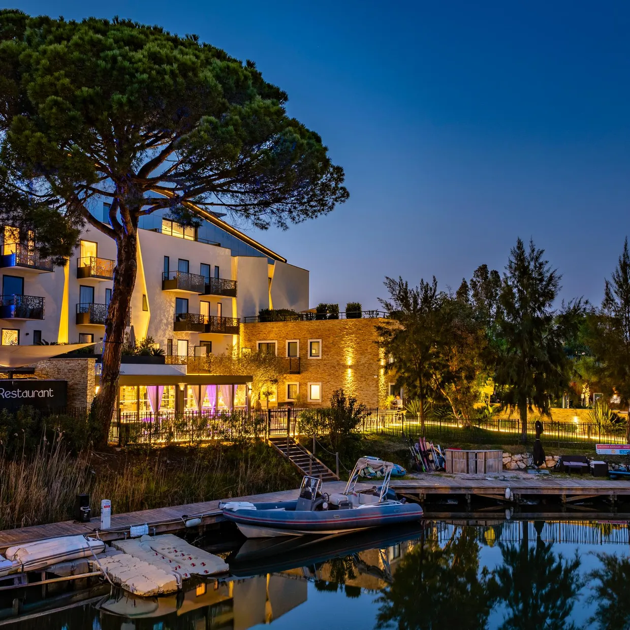 A modern building lit up with a restaurant on the ground floor, surrounded by trees and located near a body of water with moored boats.