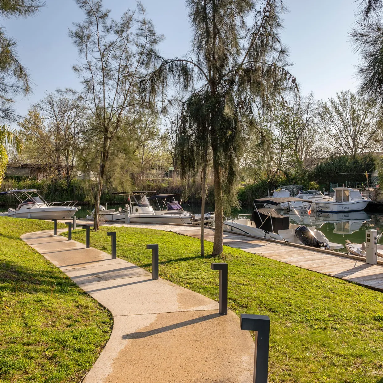 A winding path crosses a grassy area lined with trees, leading to boats moored by the water's edge.
