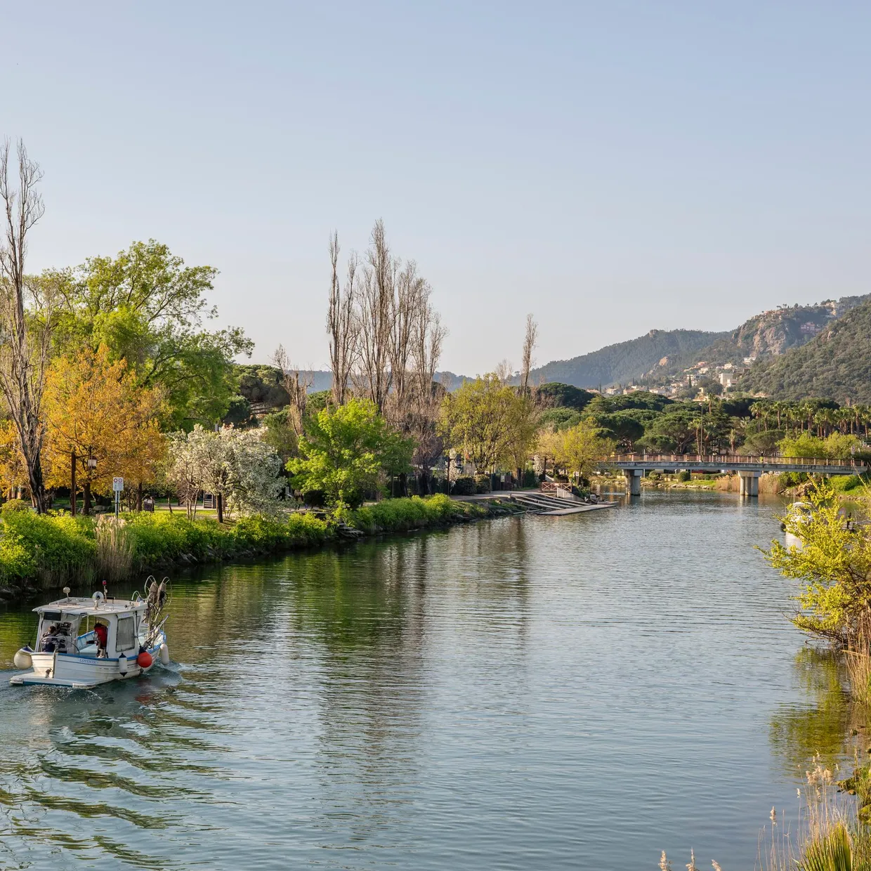 A boat sails on a river lined with trees and hills, with a bridge visible in the distance.