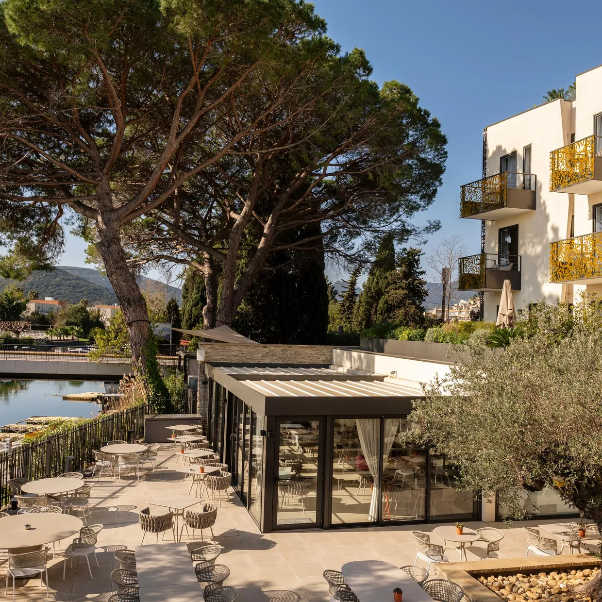 Restaurant terrace with tables and chairs near a body of water, surrounded by trees and modern buildings.
