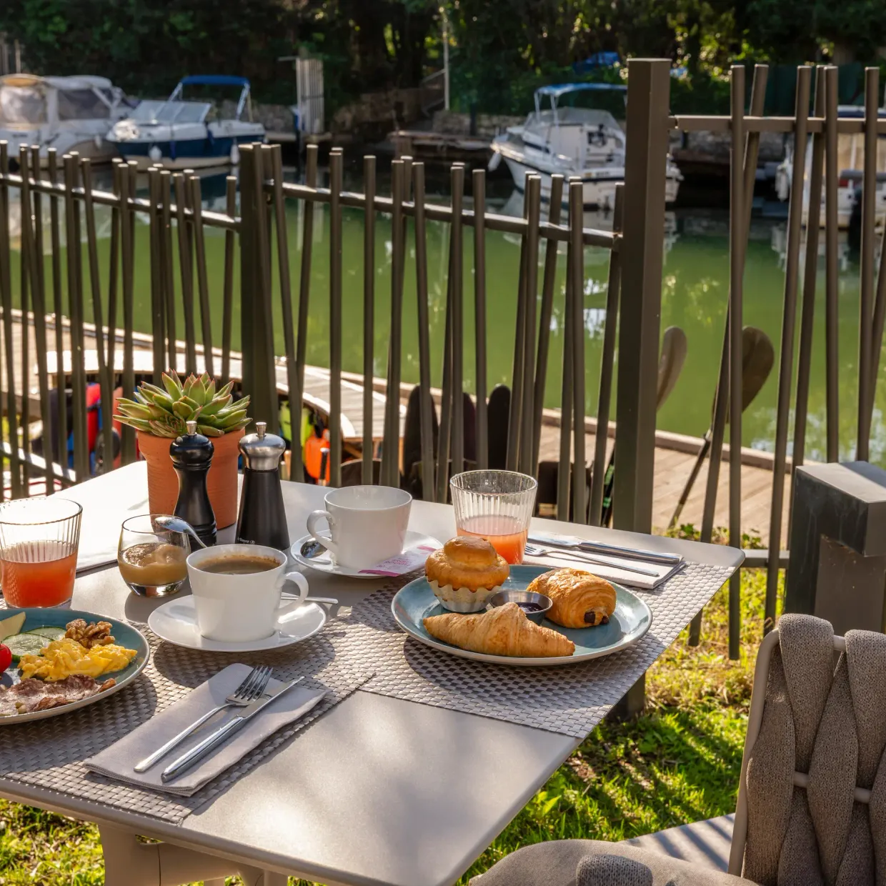 Outdoor table with breakfast consisting of croissants, bread, jam, fruit, and drinks, view of a canal with boats