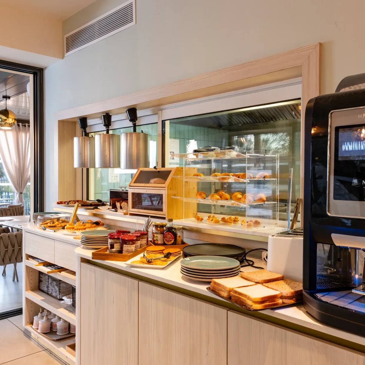 A breakfast counter with pastries, toast, jam, and a coffee machine, in a bright room with nearby tables and chairs.