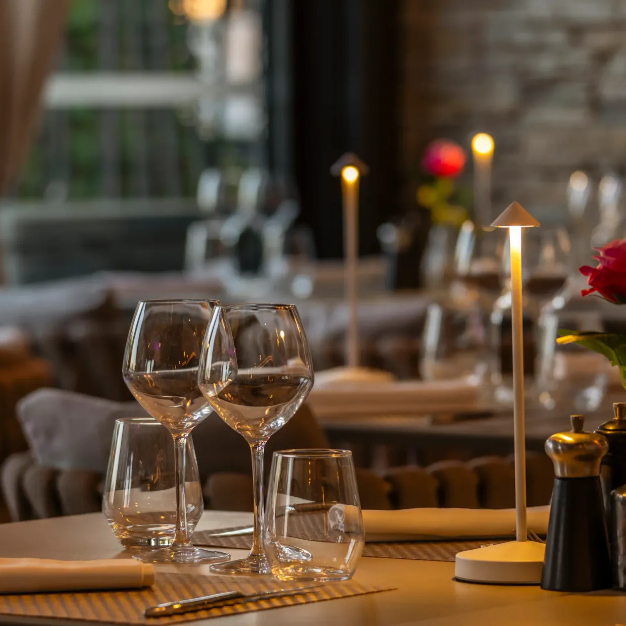 Table set with glasses, tablecloth, red rose, and candles in a dimly lit restaurant.