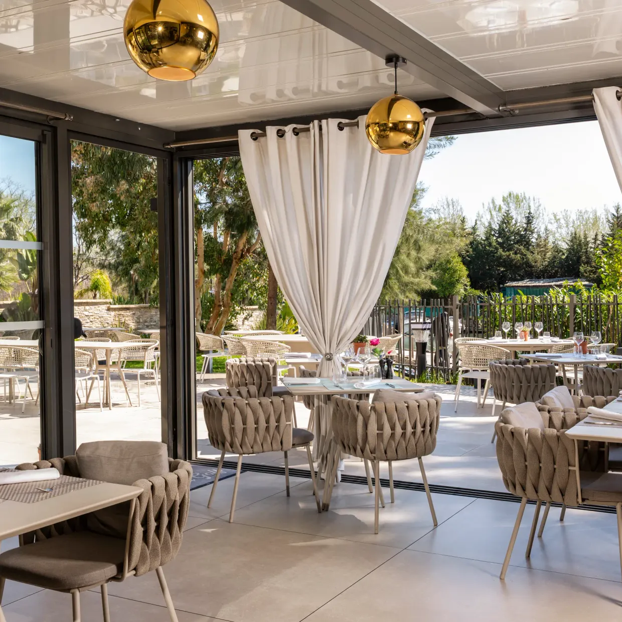 Restaurant room with tables and chairs arranged, large glass windows open onto an outdoor terrace, decorated with plants and spherical light fixtures.