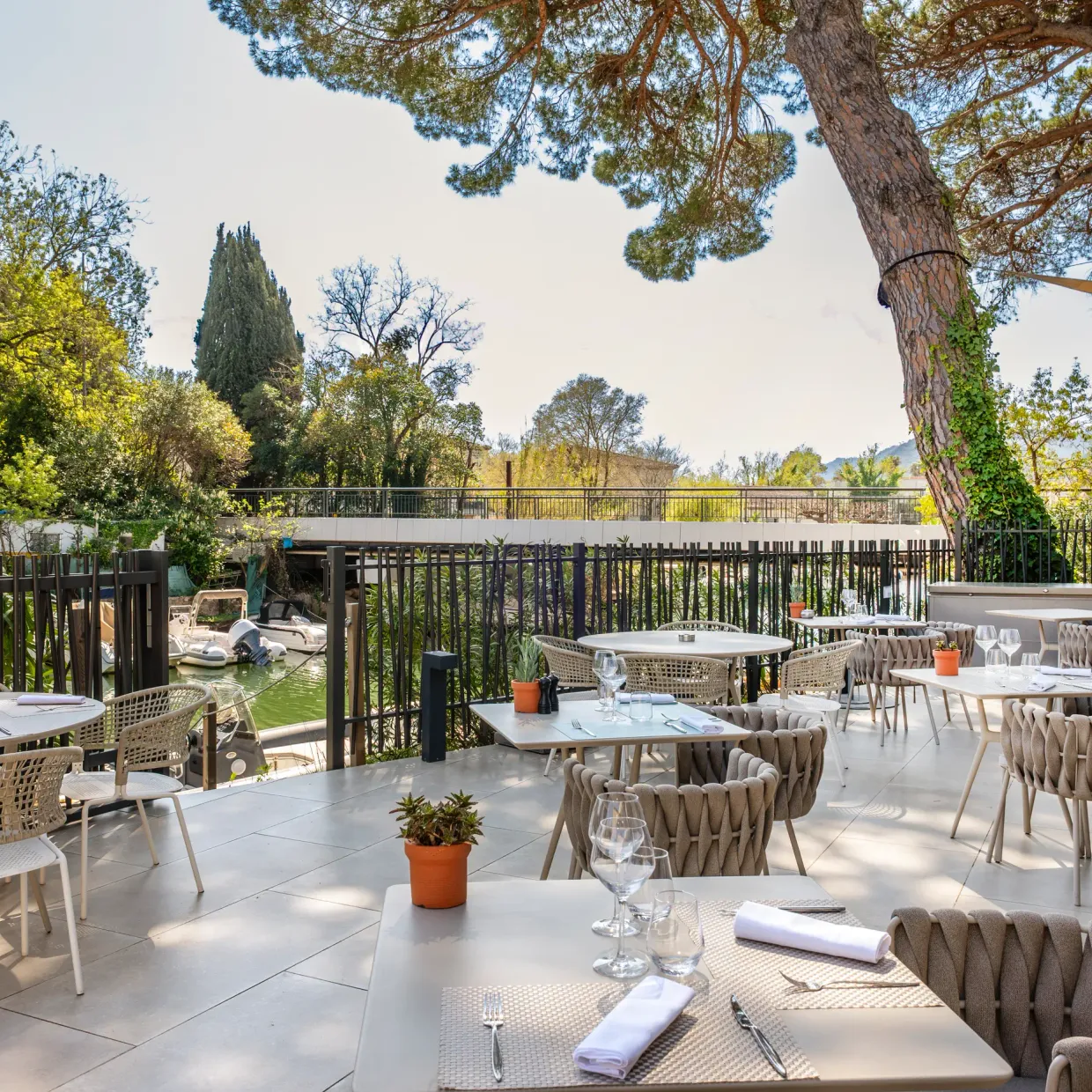 Restaurant terrace with tables and chairs arranged under a pergola, surrounded by trees and greenery.