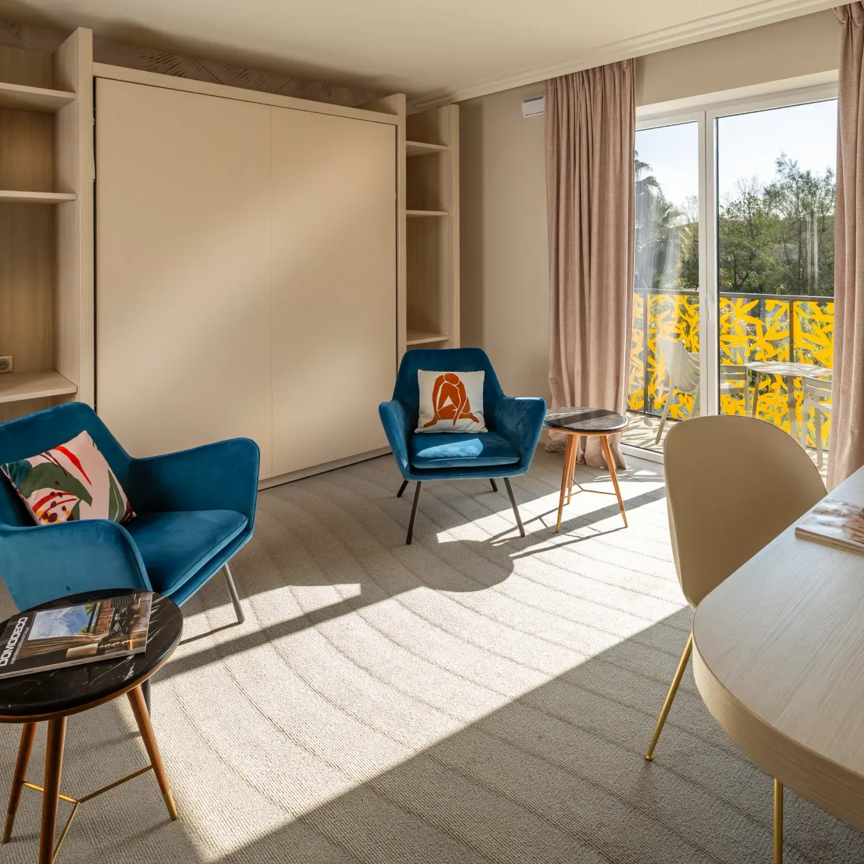 Room with two blue armchairs, a round table, a desk, shelves, and a French window opening onto a balcony with a view of trees.