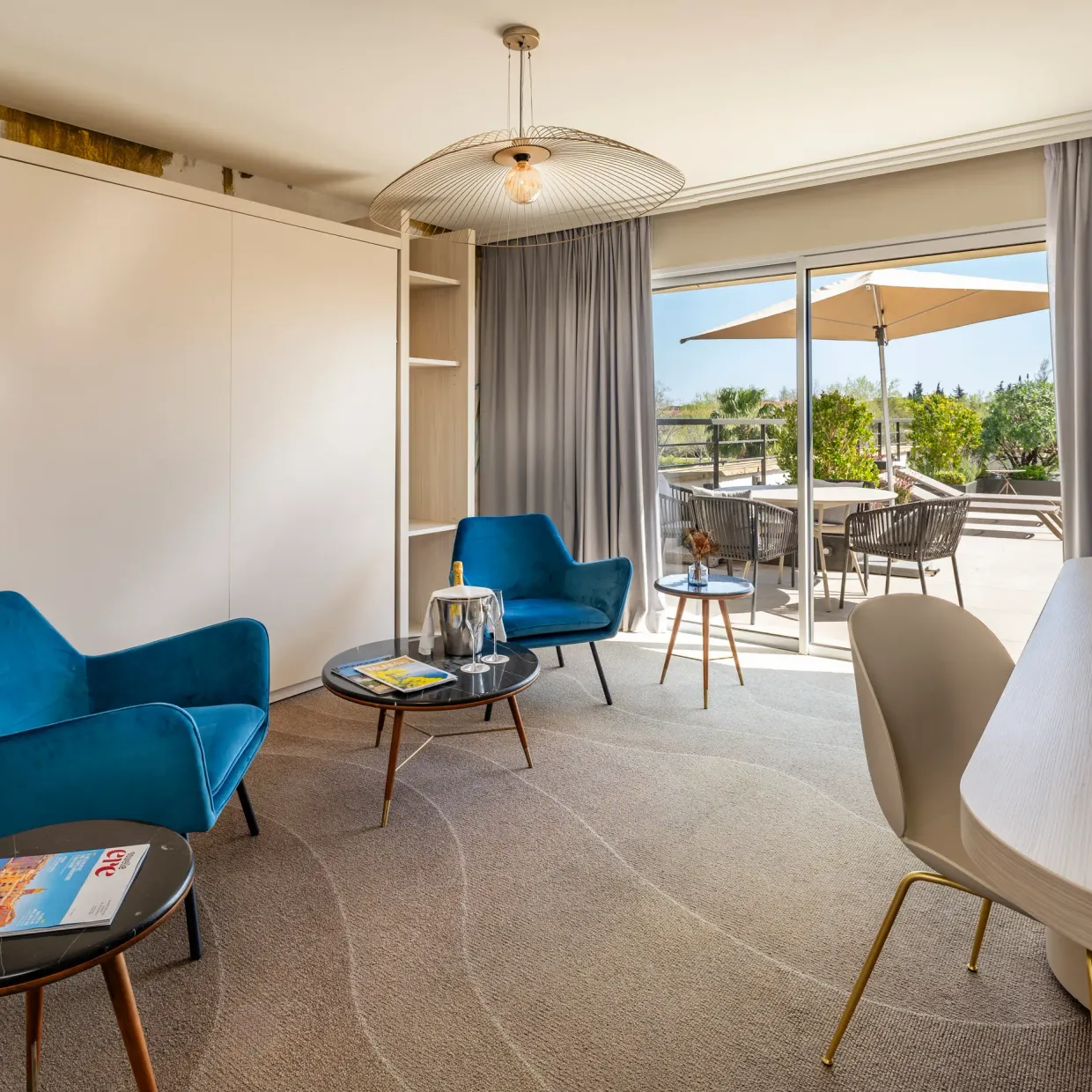 Modern living room with two blue armchairs, a coffee table, a desk, and a view of an outdoor terrace with chairs and umbrellas.