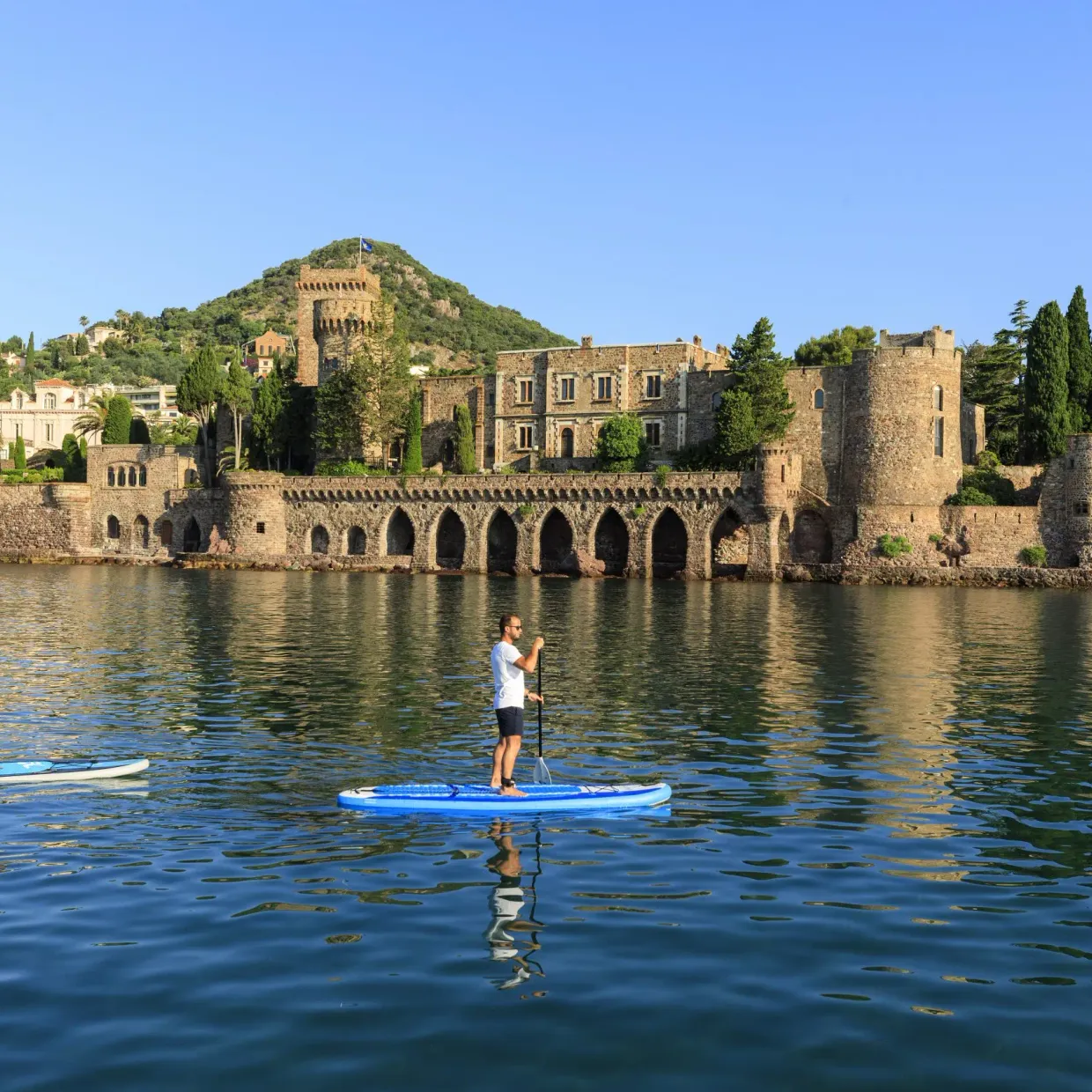 Two people are paddleboarding on a body of water in front of a medieval castle surrounded by greenery.