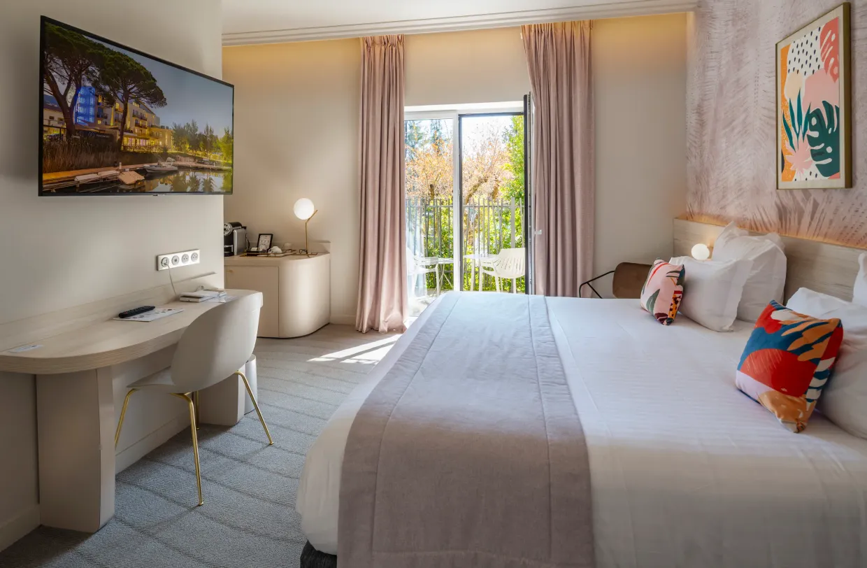 Hotel room with a bed, a desk, a television, a French window leading to a balcony, and artwork on the walls.