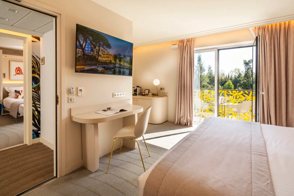 Hotel room with a bed, a desk, a television, and a French window opening onto a balcony with a view of trees.