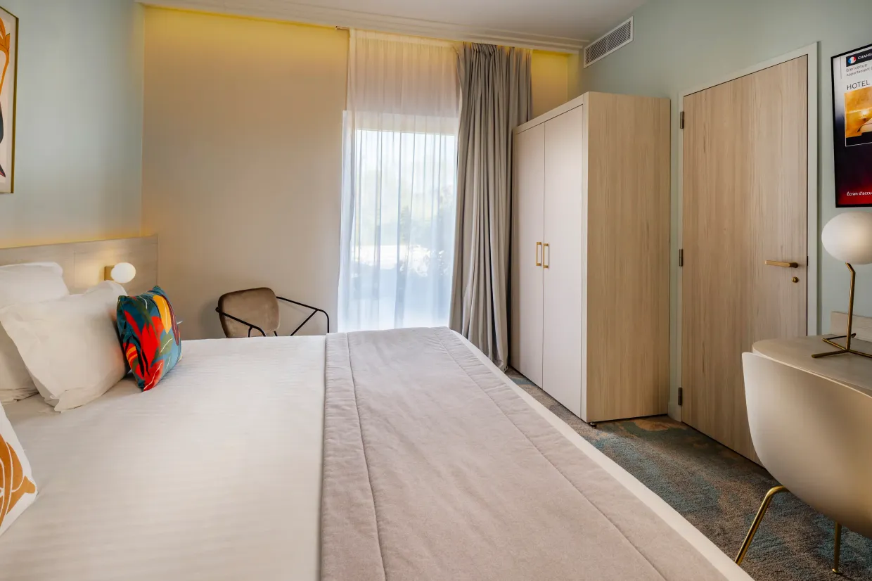 Hotel room with a double bed, a window with curtains, an armchair, a desk, and a wardrobe.