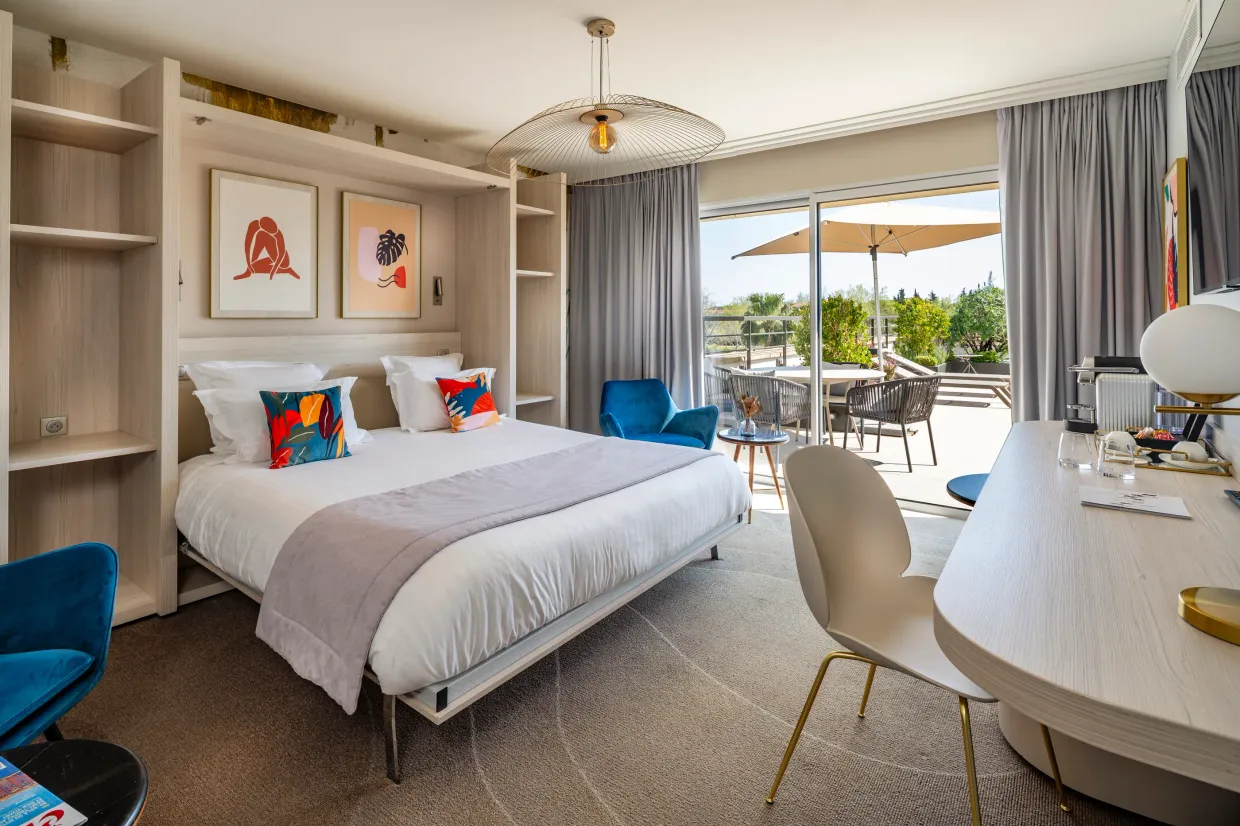 Modern hotel room with a double bed, a work desk, a blue armchair, and a view of an outdoor terrace with tables and chairs.