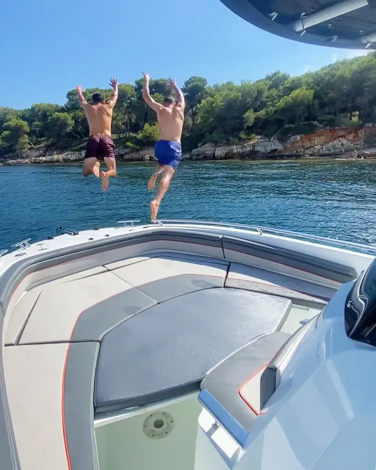 Two men jump from the back of a boat into a blue sea surrounded by vegetation and rocks.