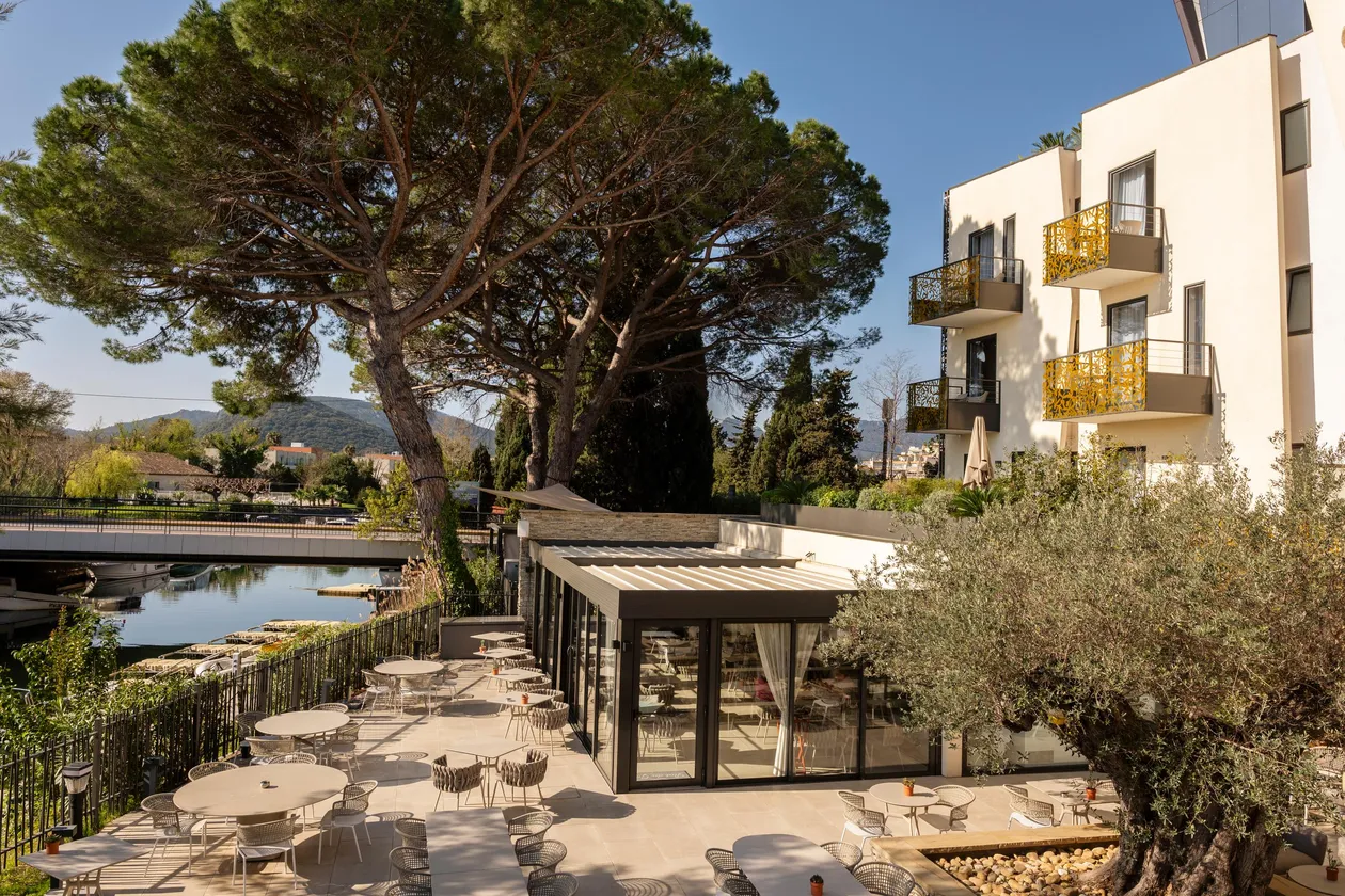 Restaurant terrace with tables and chairs near a body of water, surrounded by trees and modern buildings.