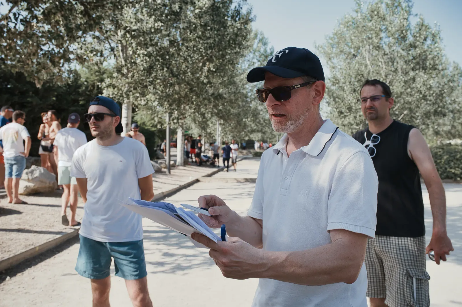 A man in a white shirt and cap reads documents outdoors, surrounded by people in summer attire.
