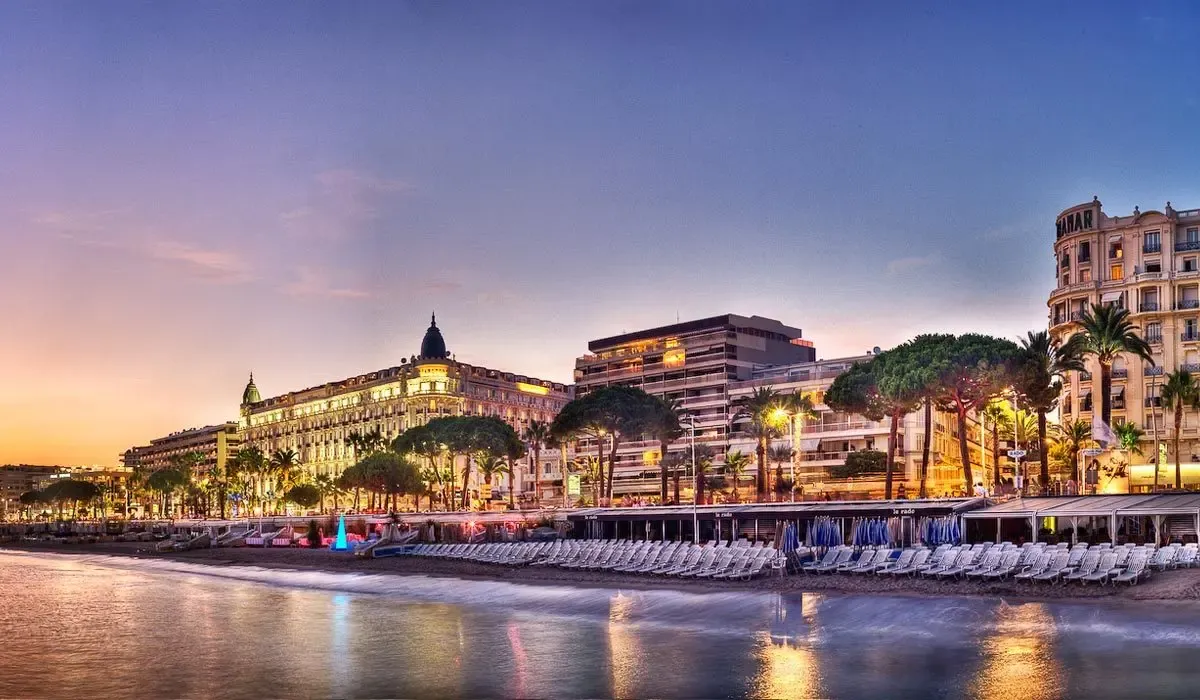 View of a coastal walkway at sunset with illuminated buildings, palm trees, and empty lounge chairs along the beach