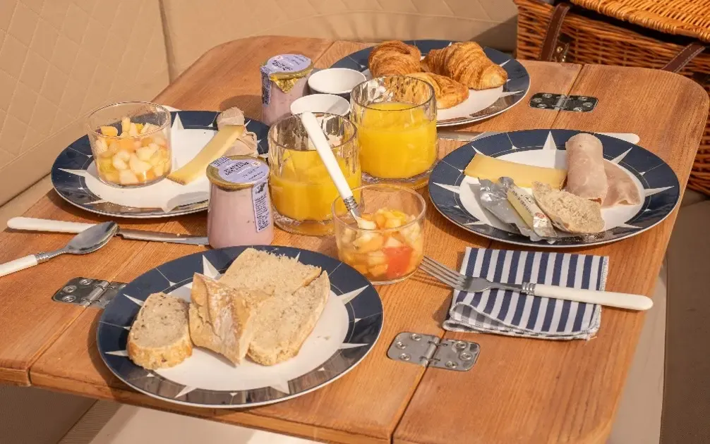 Breakfast table with dishes containing croissants, fruit, cheese, cold cuts, toast, jam, orange juice, and cutlery.