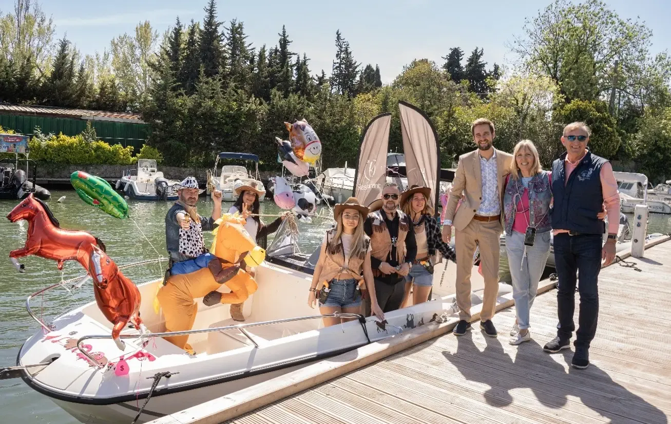 A group of people pose on a boat decorated with animal-shaped balloons, near a dock with boats in the background.