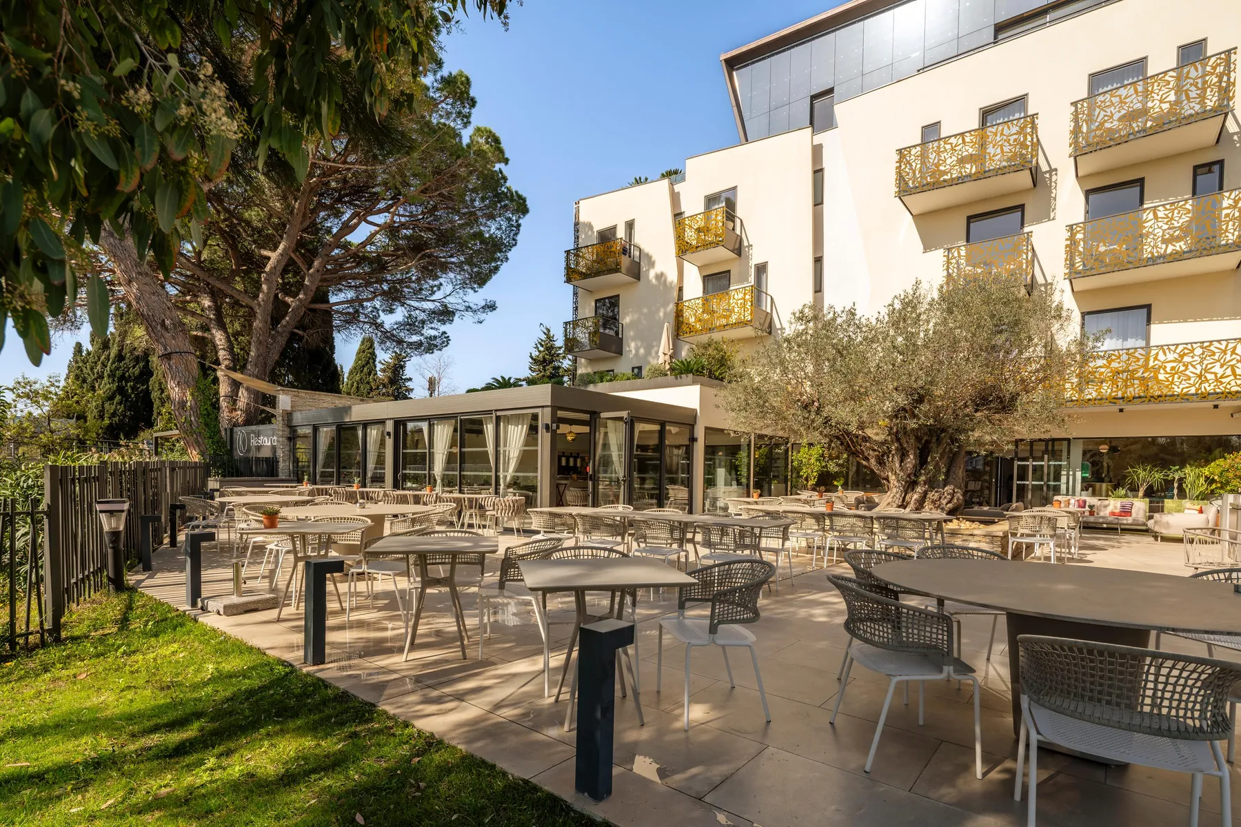 Outdoor terrace with tables and chairs, surrounded by trees and buildings with balconies adorned with geometric patterns