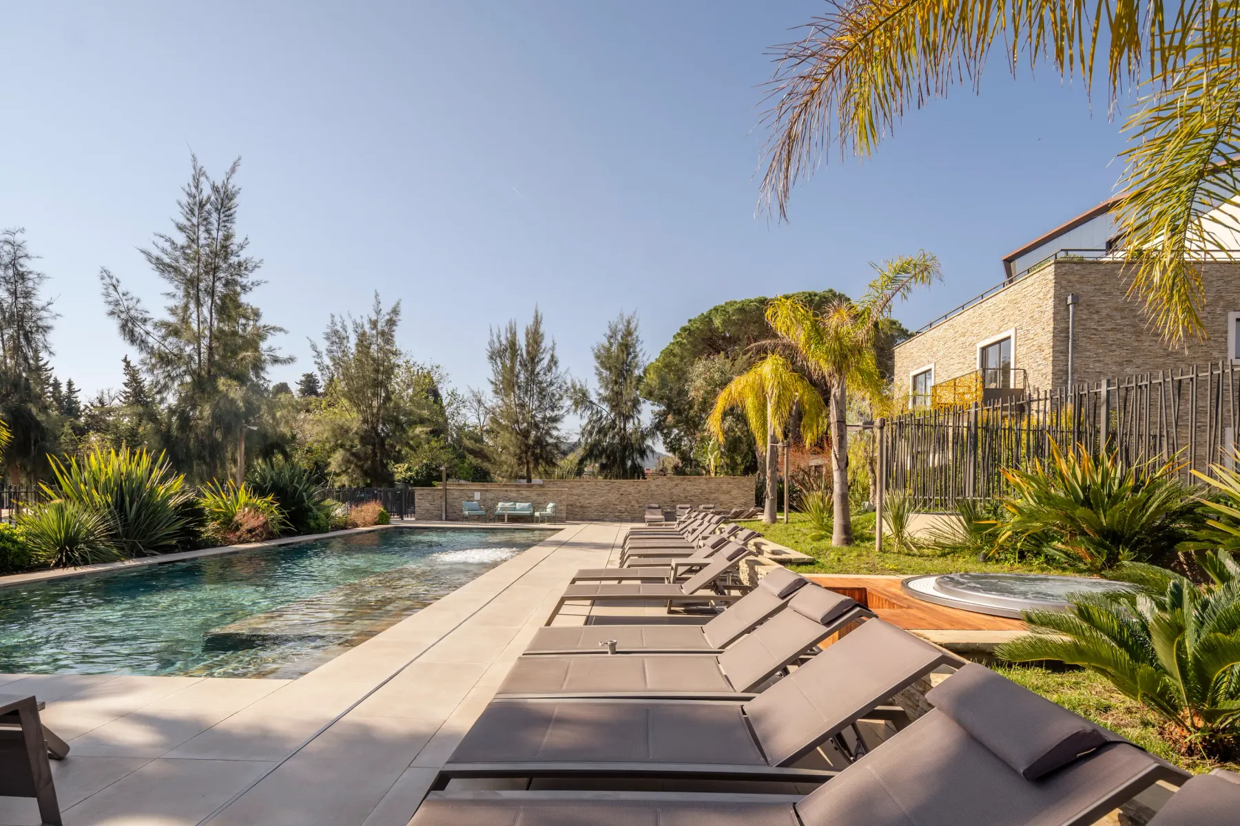 Rectangular swimming pool surrounded by sun loungers and vegetation, with a stone building in the background