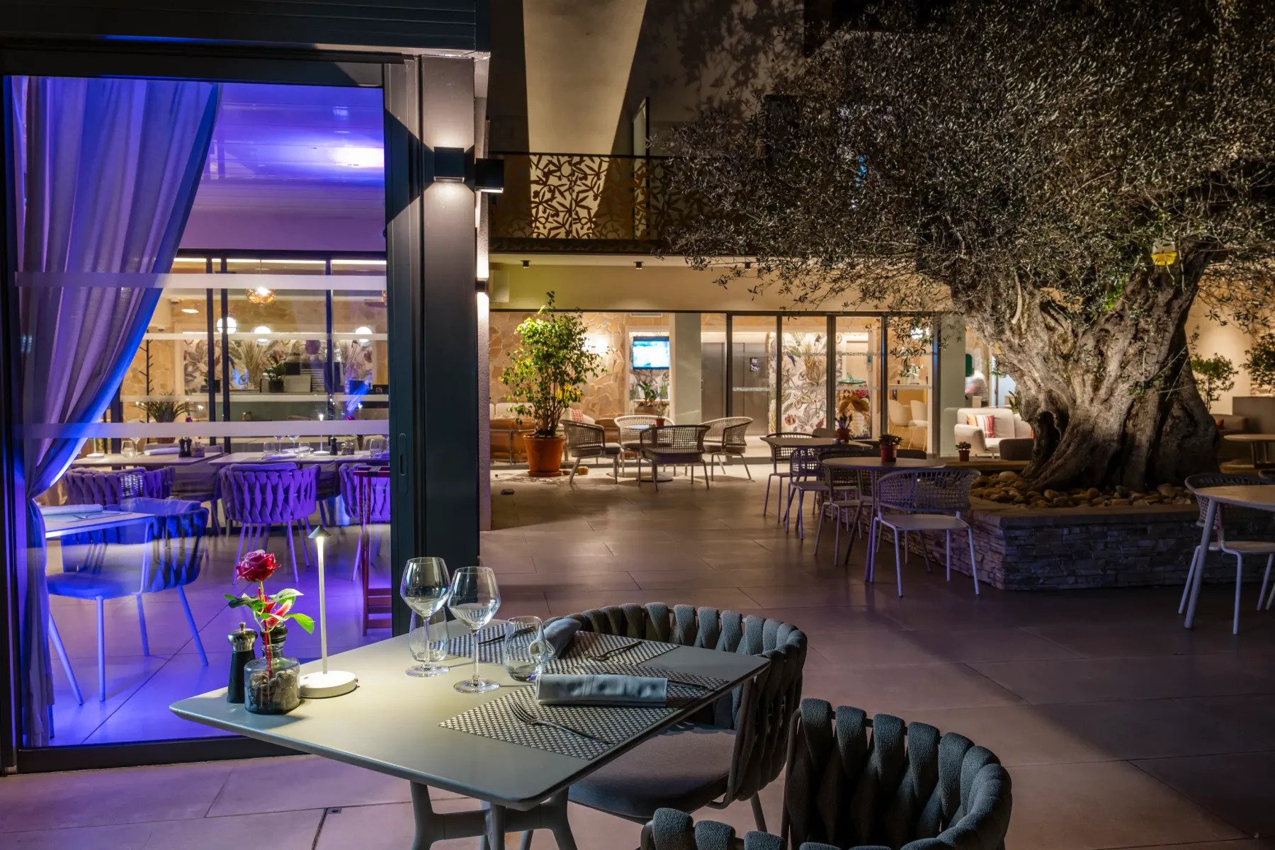 Outdoor restaurant with tables and chairs, lit by purple lights, a central tree, and a view of the indoor area