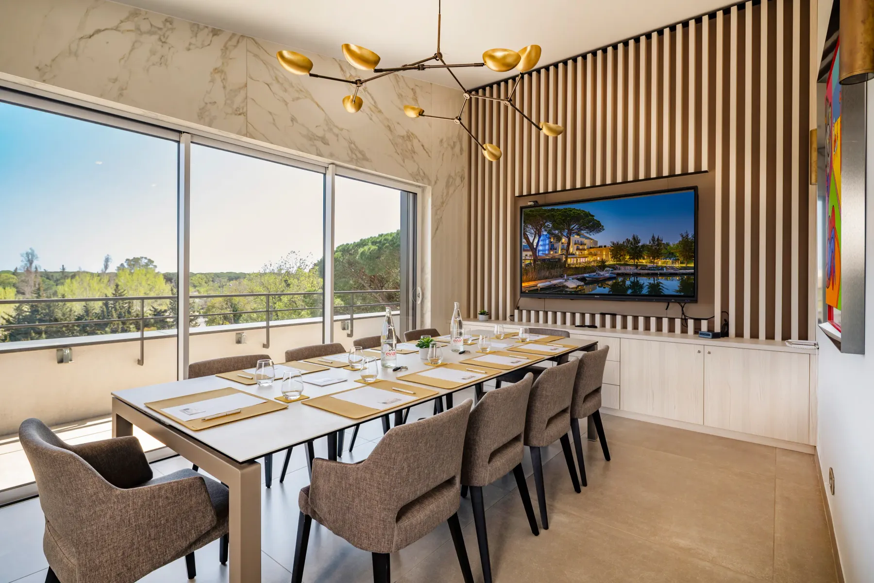 Meeting room with a table, chairs, a television displaying a landscape, and large windows offering an outdoor view.