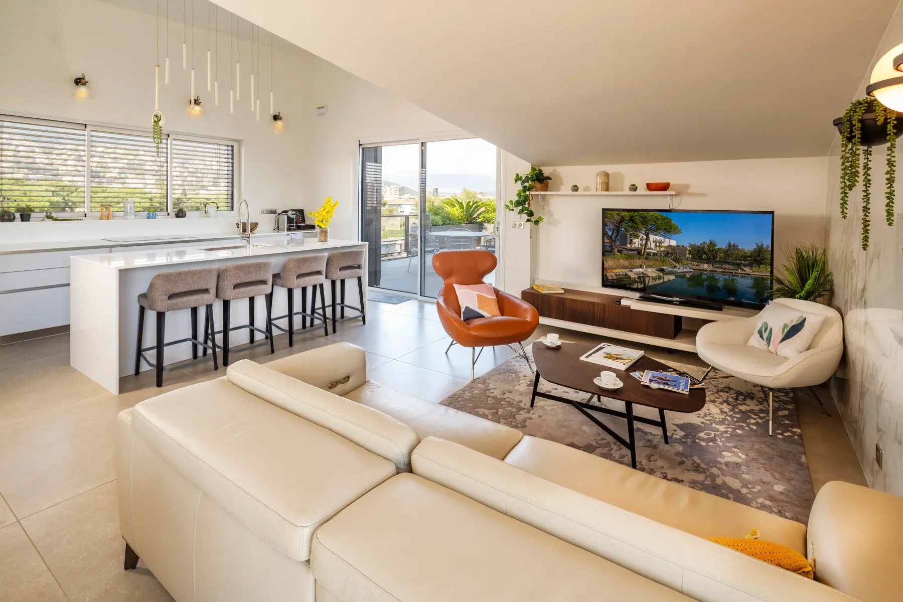 Modern living room with white sofa, armchairs, coffee table, and TV, adjacent to a kitchen with central island and stools.