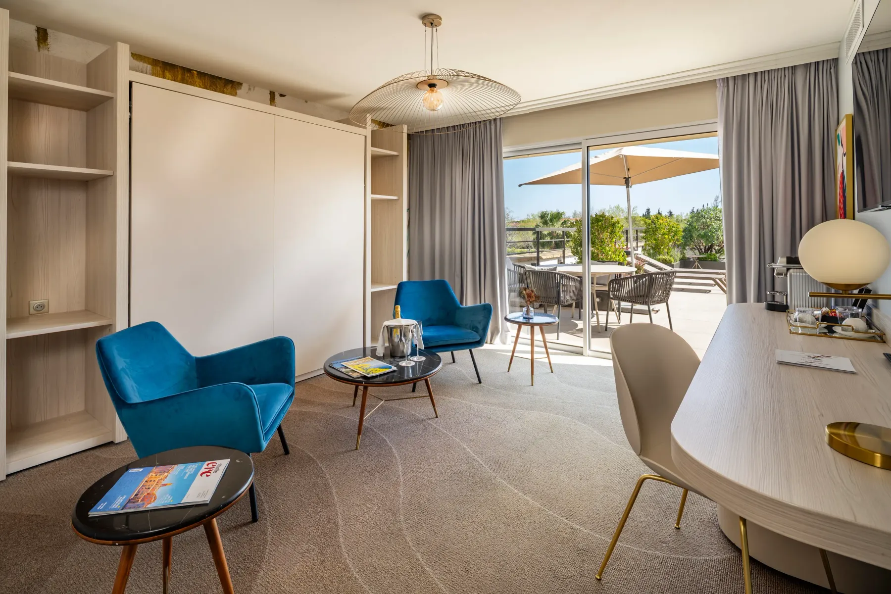 Modern living room with two blue armchairs, a coffee table, a desk, and a view of an outdoor terrace with chairs and umbrellas.