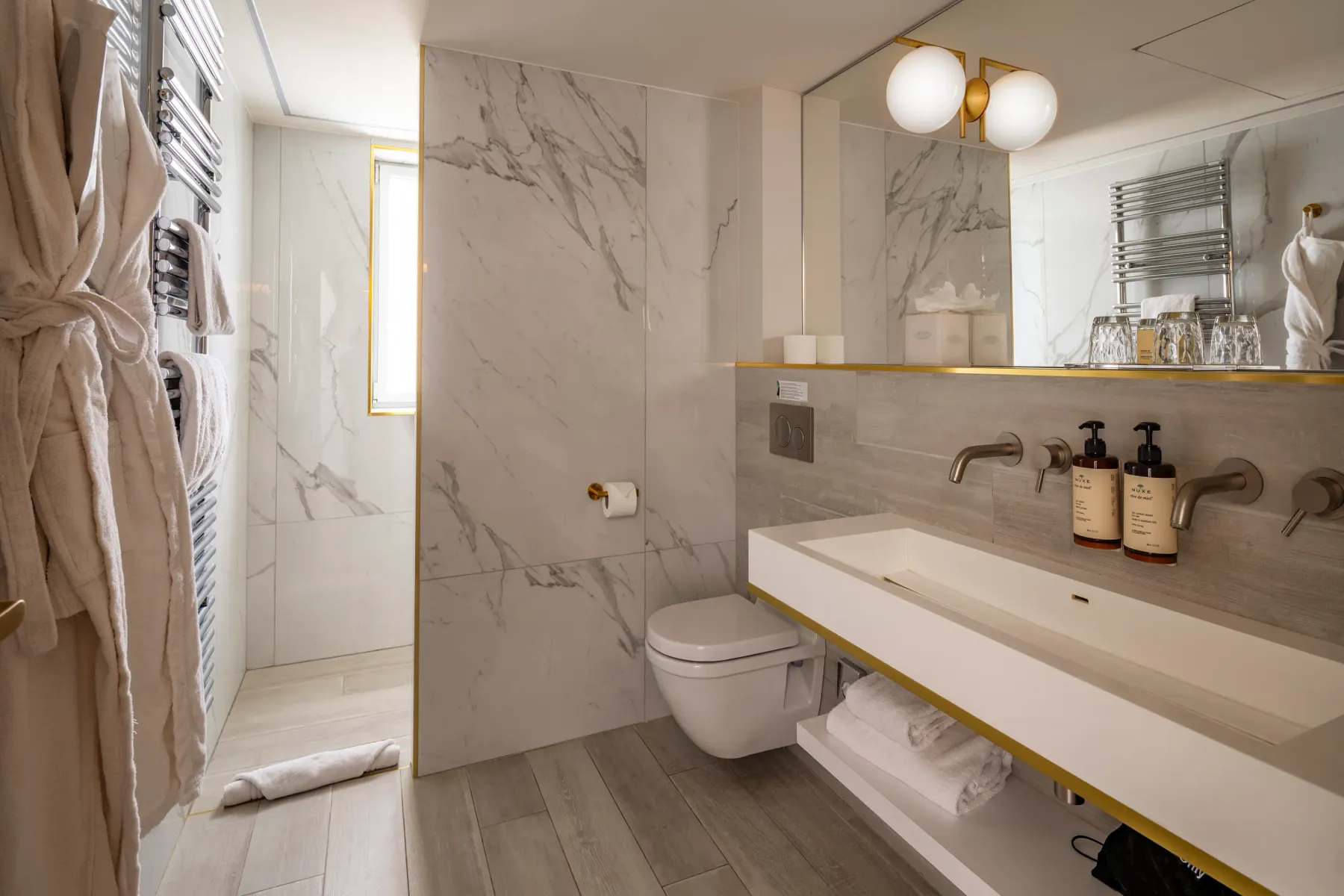 Modern bathroom with sink, toilet, mirror, and marble shelves, hanging towels, and toiletries