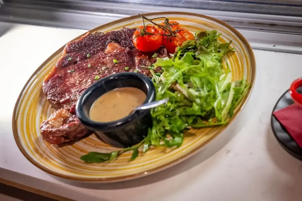An oval dish contains a piece of grilled meat, a small green salad, cherry tomatoes, and a ramekin of sauce.