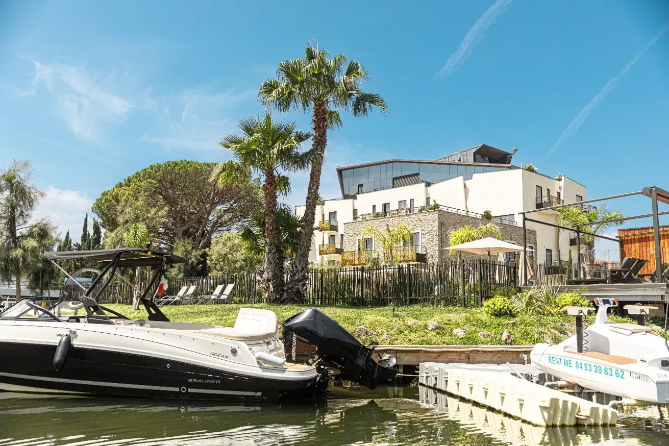 Two boats moored in front of a modern house with a garden, palm tree, and terrace by a canal under a blue sky.