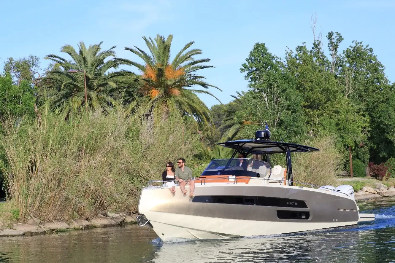 A white boat with two people on board sails on a river lined with vegetation and palm trees.