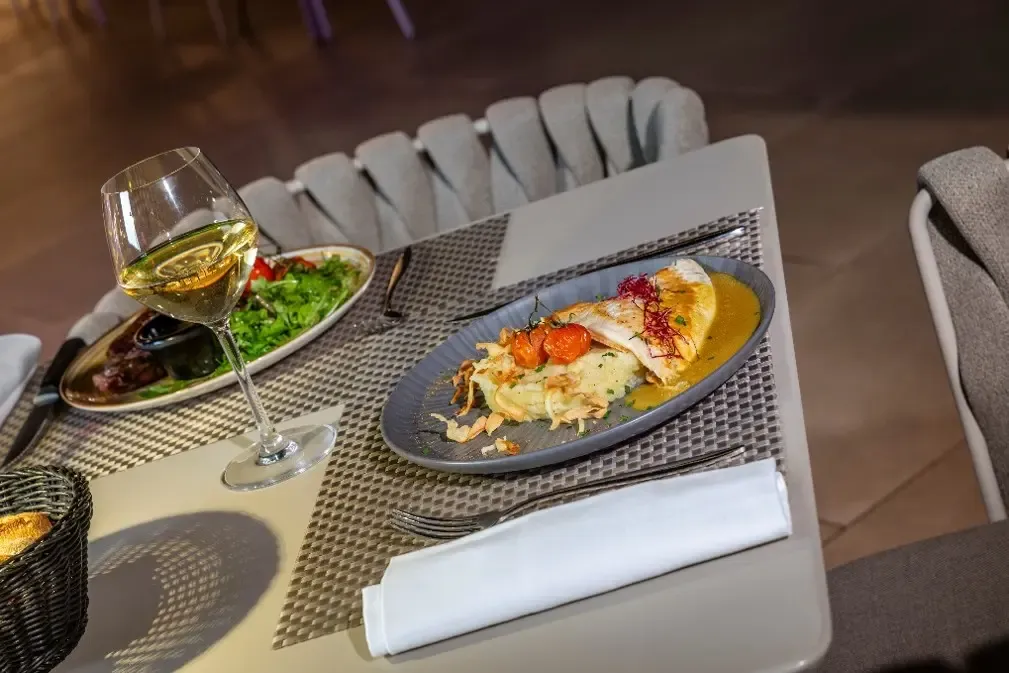 A table with a plate of grilled fish, a salad, a glass of white wine, and cutlery.