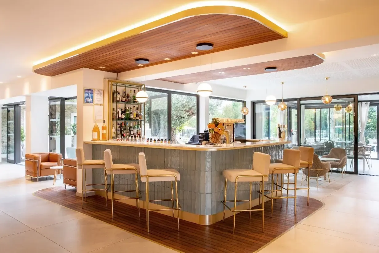 Modern bar with a gray marble counter and beige leather stools, lit by gold suspensions, surrounded by large glass windows