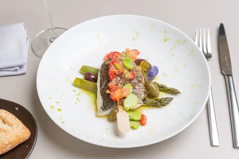 White plate with a fillet of grilled fish, asparagus, assorted vegetables, and a green sauce, served with a knife, a fork, and a wine glass.
