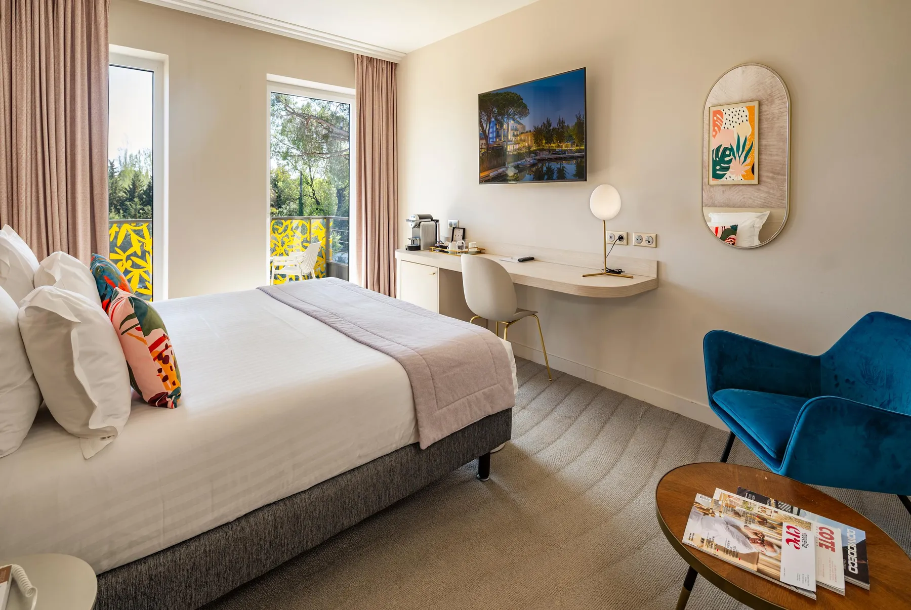 Hotel room with a bed, a work desk, a blue armchair, a television, and a view of a balcony with colorful patterns.