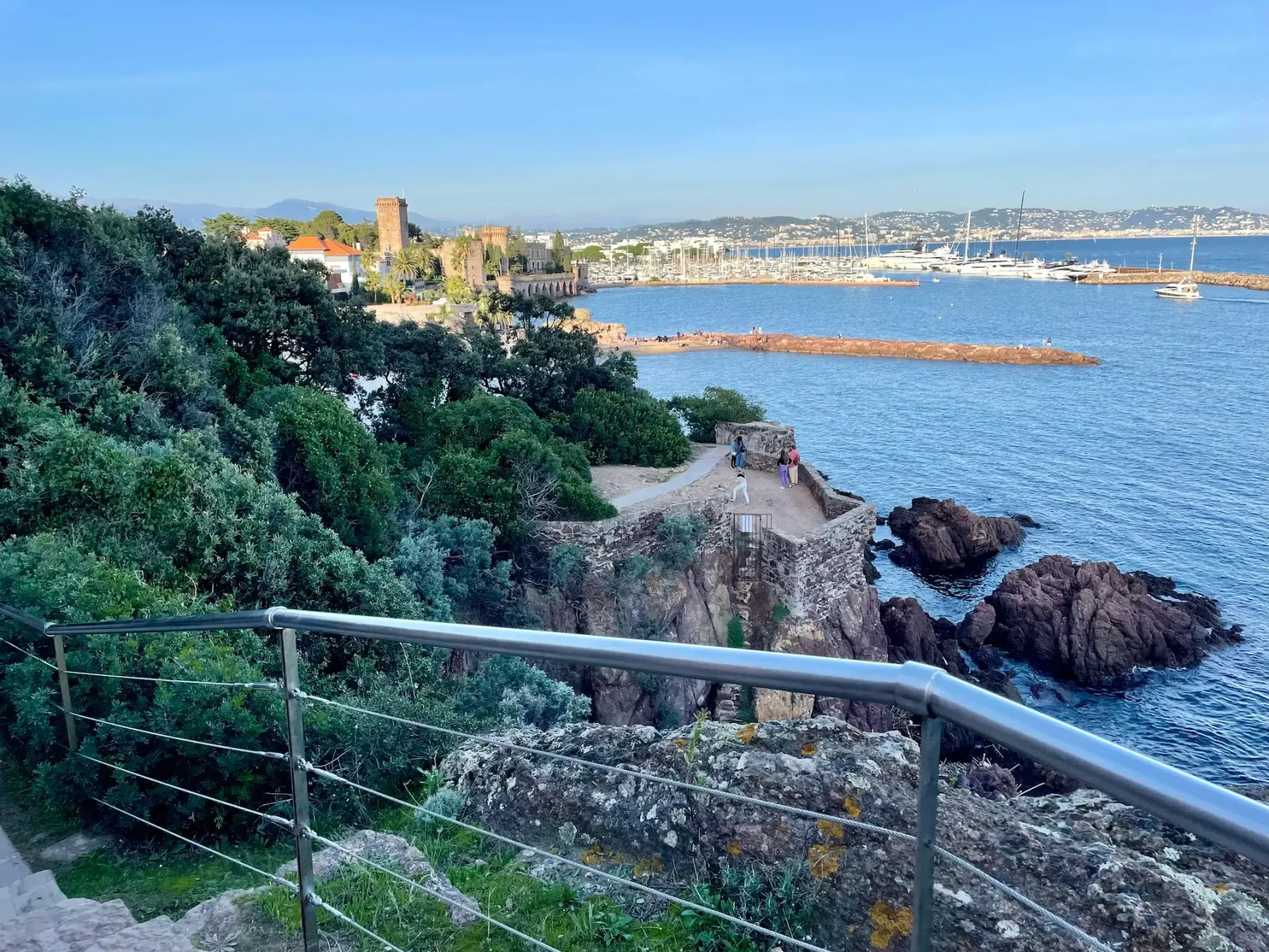 View of a marina with boats, a medieval fortress, and walkers on a coastal path