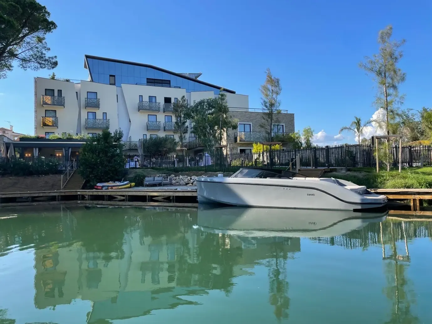 A modern building with balconies and glass windows reflects in the calm water of a canal, with a white boat moored to a wooden dock.